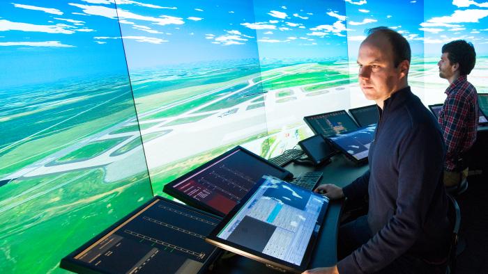 Employees in the IDEA Lab simulator standing in front of a large video display of an airport